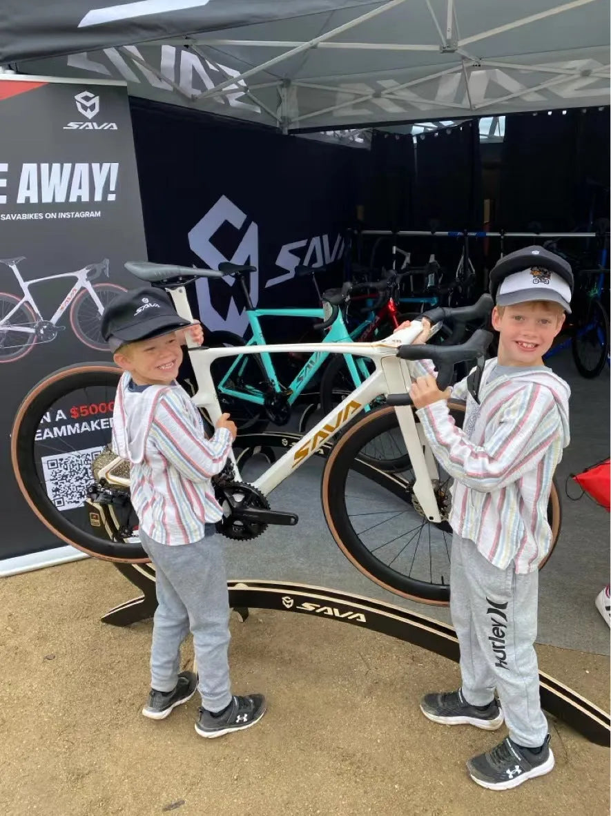 Two smiling children pose with a SAVA carbon road bike at a SAVA Carbon Bike outdoor display booth.