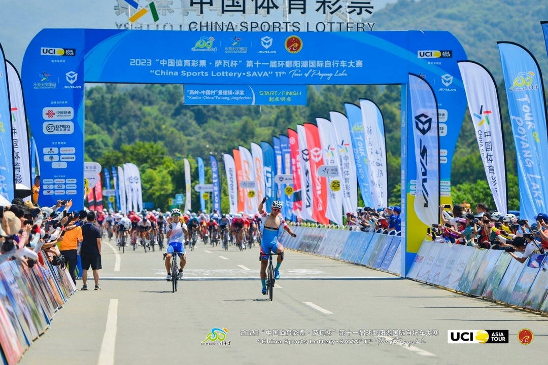 Cyclists cross finish line at SAVA Carbon Bike-sponsored road race, vibrant banners, cheering crowd