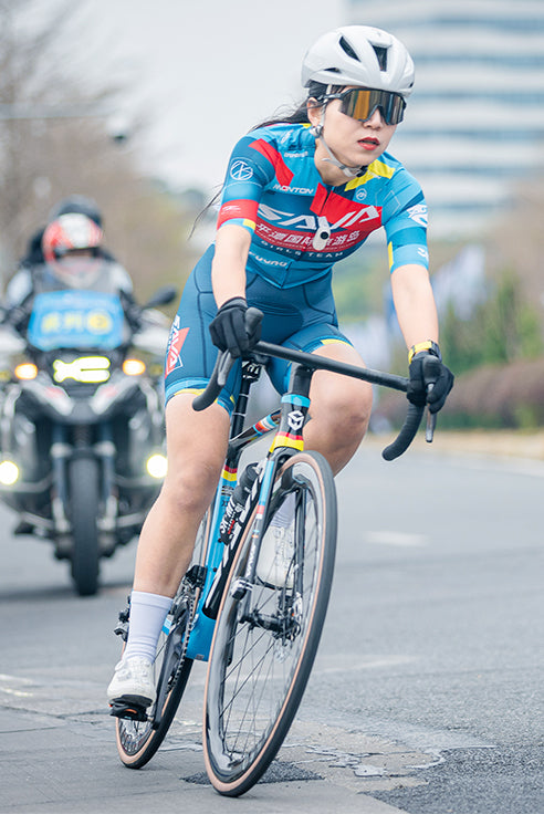 Female cyclist riding SAVA carbon road bike in race, wearing blue kit and helmet on city street